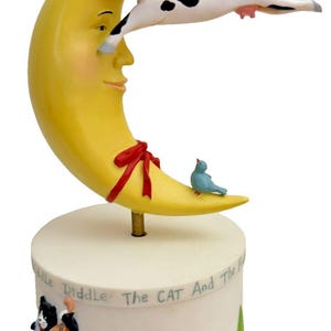 May include: A whimsical figurine featuring a yellow crescent moon with a smiling face, a red bow, and a cow jumping over it. A blue bird sits on the moon. The base has illustrations of a cat playing a cello and a dog. The text on the base reads "Diddle The CAT And The F".