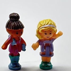 Könnte beinhalten: Zwei kleine Kunststofffiguren stehen nebeneinander. Die linke Figur hat dunkelbraunes Haar zu einem Dutt gebunden, ein magentafarbenes Oberteil und blaue Stiefel. Die rechte Figur hat blondes Haar mit einem violetten Stirnband und grüne Stiefel.
