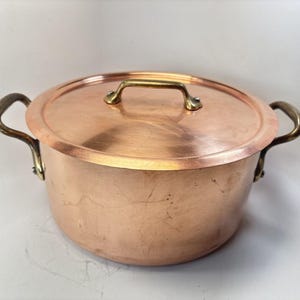 May include: A copper cooking pot with a lid and brass handles. The pot has two side handles and a small handle on the lid. The copper has a shiny, reflective surface.