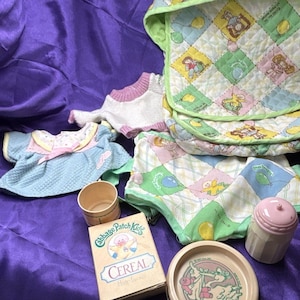 May include: A collection of vintage Cabbage Patch Kids items, including clothing, a bib, a cereal box, a small bowl, and a container with a pink lid. The items are arranged on a purple fabric surface.