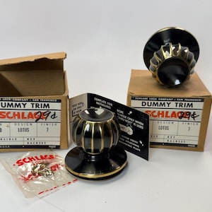May include: Two Schlage dummy trim door knobs with a black and brass finish, featuring a fluted lotus design. Includes two cardboard boxes labeled with product details, a black installation template, and a small plastic bag containing metal screws.