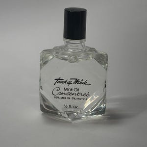 Könnte beinhalten: Eine durchsichtige Glasflasche mit schwarzem Verschluss, die Touch of Mink Concentrée Nerzöl enthält. Das Etikett gibt 95% Mink Oil, 5% vitamin E und 1/2 fl. oz. an. Die Flasche hat eine facettierte, geometrische Form mit einem schmalen Hals.