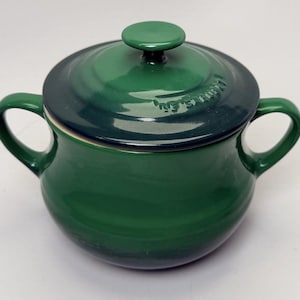 May include: A green enameled cast iron pot with a matching lid and two loop handles. The lid has a small, round knob and the brand name is embossed on the lid. The pot is a deep green color.