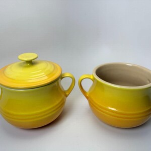 May include: Two yellow ceramic bowls with handles. One bowl has a lid with a small knob. The bowls have a gradient color, transitioning from a darker yellow at the base to a lighter yellow at the top. The interior of the open bowl is beige.
