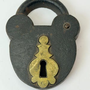 May include: Antique black padlock with a brass keyhole cover. The lock has a rounded top and a keyhole cover with an ornate design. The metal has a weathered appearance, suggesting age and use. A vintage security item.