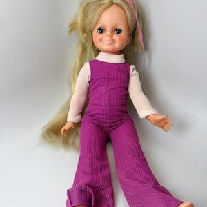 May include: A vintage doll with long blonde hair and blue eyes. The doll wears a pink ribbon in its hair, a white long-sleeved shirt, and a ribbed magenta jumpsuit. The doll's arms and legs are outstretched, and it has a smiling expression.