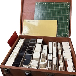 May include: Vintage brown suitcase open to reveal a collection of miniature kitchen furniture pieces in white and black. Includes a green cutting mat and a yellow card. The suitcase is made of brown leather.