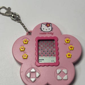 Könnte beinhalten: Ein rosa, blumenförmiges elektronisches Hello-Kitty-Handheld-Spiel mit einem digitalen Bildschirm, der ein Schachbrettmuster anzeigt. Es verfügt über gelbe blumenförmige Tasten mit roten Herzen, weiße Richtungspfeile und einen silbernen Metallschlüsselanhänger.