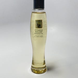 Könnte beinhalten: Eine klare, schlanke Glasflasche mit hellgelbem Massageöl und einem schwarzen Verschluss. Das Etikett trägt die Aufschrift: eucalyptus & spearmint oils, STRESS RELIEF MASSAGE OIL, infused with St. John's Wort. Die Flasche hat eine geschwungene Form.