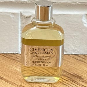 May include: A small, clear glass bottle of Givenchy Gentleman after-shave lotion. The bottle has a silver cap and a label with the product name and details. The bottle contains 1 fl oz, or 30 ml, of after-shave lotion.