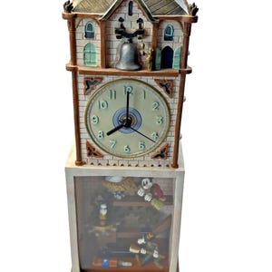 May include: A decorative clock featuring a bell tower design with a clock face and animated characters. The clock has a beige brick facade, a gray roof, and a wooden base. The clock face has black numerals and hands.