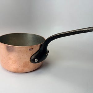 May include: A copper saucepan with a black handle. The pan has a rounded shape and a shiny interior. The handle is riveted to the side of the pan. The words "MADE IN FRANCE" are stamped on the side of the pan.