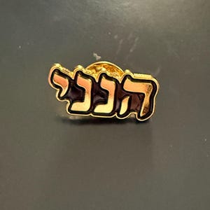 Puede incluir: Pin dorado con texto hebreo en una fuente estilizada. El pin tiene un fondo oscuro con bordes dorados, y el texto parece ser de color marrón claro. El pin es probablemente una joya o un accesorio.
