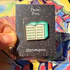 Könnte beinhalten: Ein Ansteck-Pin mit einem türkisblauen Buch mit goldenen Rändern und hebräischen Texten. Der Pin befindet sich auf einer schwarzen Karte mit dem Text "Pnimi Pins" und dem Instagram-Handle "@pnimipins". Der Pin ist in einer transparenten Plastikhülle.