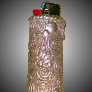 May include: A decorative lighter with a textured, iridescent surface. The lighter is covered in swirling patterns and glitter, with a red button and a green metal top. The design is a unique, artistic take on a common item.