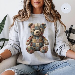 May include: Ash-colored sweatshirt featuring a patchwork teddy bear design. The bear has a variety of colors, including blue, red, and tan. The sweatshirt is a light gray color and has a crew neck. The word "Ash" is printed on the upper right corner.