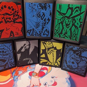 May include: A collection of seven black boxes, each featuring a different character illustration in vibrant colors. The illustrations include red, blue, green, yellow, and gray designs. The boxes appear to be made of a textured material.
