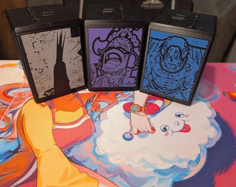 May include: Three black card storage boxes with unique graphic designs. One features a gray silhouette of a building, another a purple illustration of a character, and the third a blue illustration. The boxes are on a colorful playmat.