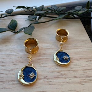 Puede incluir: Un par de dilatadores de oreja dorados con un dije colgante. El dije presenta una luna creciente con una cara y un diseño de estrella sobre un fondo azul oscuro. Los dilatadores se muestran sobre una superficie de madera.
