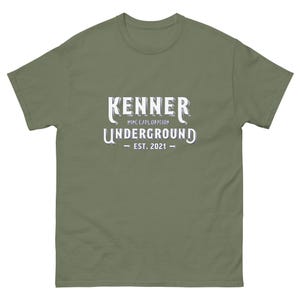 May include: Olive green t-shirt with the text "KENNER MINE EXPLORATION UNDERGROUND EST. 2021" in white. The shirt has short sleeves and a crew neck.