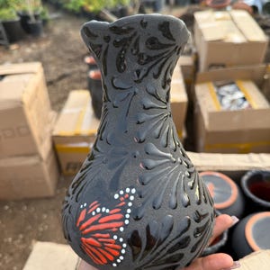 May include: A dark gray ceramic vase with a butterfly design. The vase features a textured surface with embossed floral patterns and a painted butterfly in red, white, and black. The vase is held in a hand.