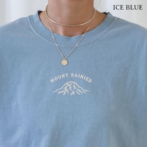 以下が含まれることがあります： 「MOUNT RAINIER」の文字が白で刺繍された、山岳グラフィックの入ったアイスブルーのTシャツ。ペンダントとチョーカーが付いたゴールドカラーのネックレスが着用されています。「ICE BLUE」の文字が右上隅にあります。