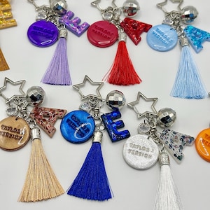 May include: A collection of keychains with star-shaped charms, disco ball accents, and colorful tassels. Each keychain features a letter or a circular pendant with the text "Taylor's Version" in a variety of colors, including purple, red, and blue.