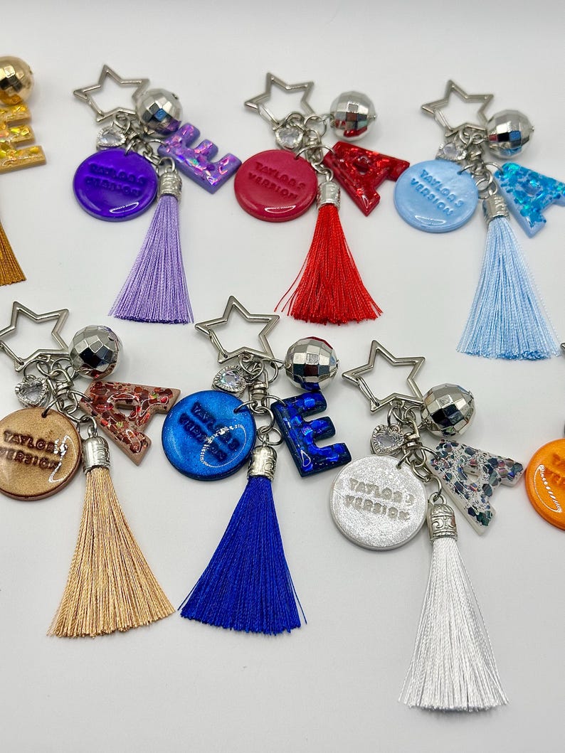 May include: A collection of keychains with star-shaped charms, disco ball accents, and colorful tassels. Each keychain features a letter or a circular pendant with the text "Taylor's Version" in a variety of colors, including purple, red, and blue.