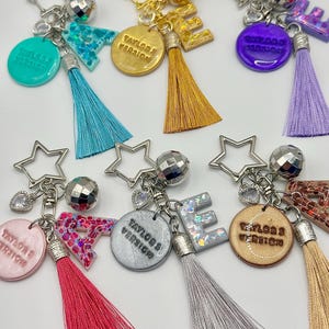 May include: A collection of keychains with star-shaped charms, disco ball accents, and colorful tassels. Each keychain features a letter or a circular pendant with the text "Taylor's Version". The keychains come in various colors, including teal, gold, purple, pink, silver, and tan.