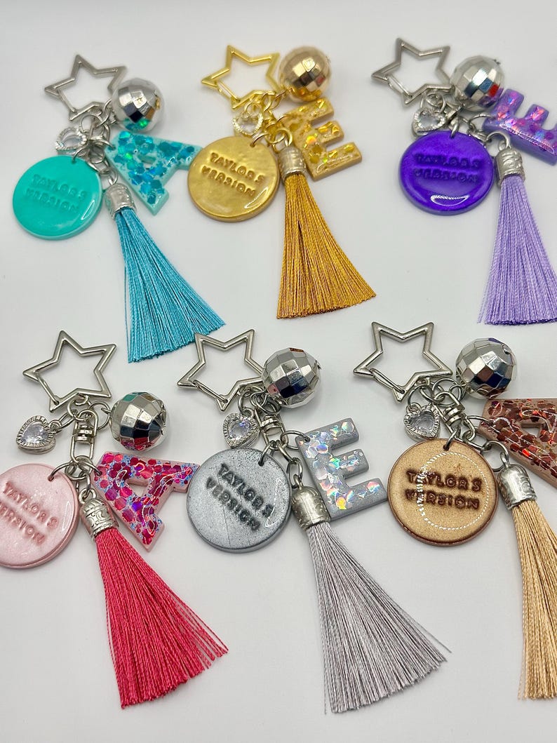 May include: A collection of keychains with star-shaped charms, disco ball accents, and colorful tassels. Each keychain features a letter or a circular pendant with the text "Taylor's Version". The keychains come in various colors, including teal, gold, purple, pink, silver, and tan.