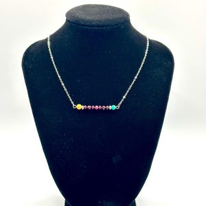 Puede incluir: Un collar de cadena plateada con un colgante de barra horizontal compuesto por una cuenta amarilla, una serie de pequeñas cuentas facetadas de color rojo oscuro y una cuenta turquesa, separadas por pequeños espaciadores plateados.
