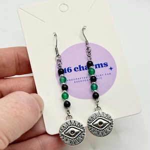 Könnte beinhalten: Ohrringe mit silbernen Augenanhängern und einem Perlendesign. Die Ohrringe haben abwechselnd schwarze und grüne Perlen. Auf der Karte steht der Text "16 charms".