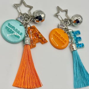 May include: Two keychains with star and disco ball charms. One keychain has a teal circle with "Taylor's Version" text, an orange letter "A", and an orange tassel. The other has an orange circle with "Taylor's Version" text, a blue letter "E", and a blue tassel.