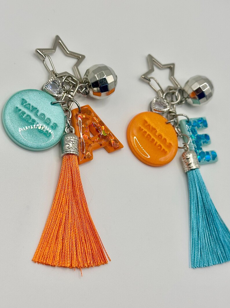 May include: Two keychains with star and disco ball charms. One keychain has a teal circle with "Taylor's Version" text, an orange letter "A", and an orange tassel. The other has an orange circle with "Taylor's Version" text, a blue letter "E", and a blue tassel.