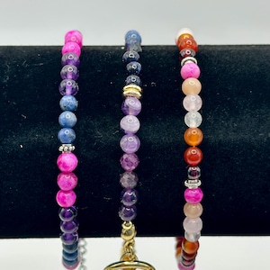 May include: Three beaded bracelets in shades of pink, purple, blue, orange, and white. The bracelets are made with round gemstone beads and feature silver and gold-coloured accents. One bracelet has a toggle clasp.