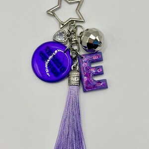May include: A keychain with a star-shaped charm, a faceted silver ball, and a purple letter "E". The keychain also features a purple tassel and a round purple charm with white text. The keychain is made of metal and resin.
