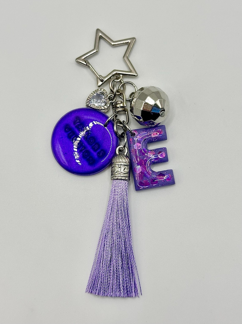 May include: A keychain with a star-shaped charm, a faceted silver ball, and a purple letter "E". The keychain also features a purple tassel and a round purple charm with white text. The keychain is made of metal and resin.