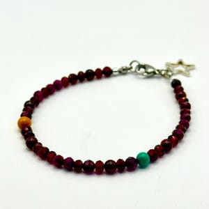 Puede incluir: Una pulsera de cuentas con pequeñas cuentas facetadas de color rojo granate oscuro, una cuenta amarilla lisa y una cuenta turquesa lisa. La pulsera incluye un cierre de mosquetón de metal plateado y un pequeño dije de estrella plateado.