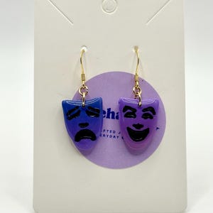 Op de afbeelding: Goudkleurige oorhangers met dramamaskers. Eén masker is blauw met een verdrietig gezicht, de andere is paars met een blij gezicht. De oorbellen worden gepresenteerd op een witte kaart met een paarse cirkel.