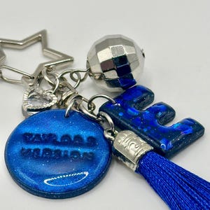 May include: A blue and silver keychain with a star, heart, and disco ball charm. The keychain features a blue letter "E" and a round charm with the text "Taylor's Version". A blue tassel hangs from the letter.