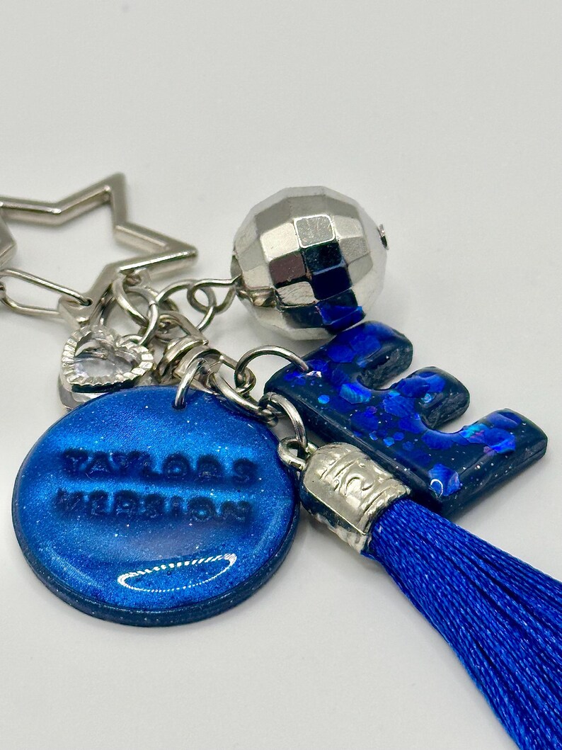 May include: A blue and silver keychain with a star, heart, and disco ball charm. The keychain features a blue letter "E" and a round charm with the text "Taylor's Version". A blue tassel hangs from the letter.