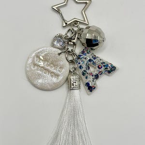 May include: A silver keychain with multiple charms. The charms include a star, a faceted ball, a heart, a letter "A" with a blue and white pattern, a round charm with text, and a white tassel.
