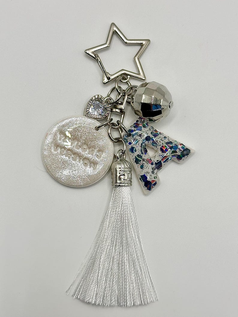 May include: A silver keychain with multiple charms. The charms include a star, a faceted ball, a heart, a letter "A" with a blue and white pattern, a round charm with text, and a white tassel.