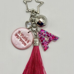 May include: A keychain with a silver star-shaped clasp, a disco ball charm, a heart charm, a pink circular charm with text, a pink glittery letter "A", and a long pink tassel. The keychain is a decorative accessory.