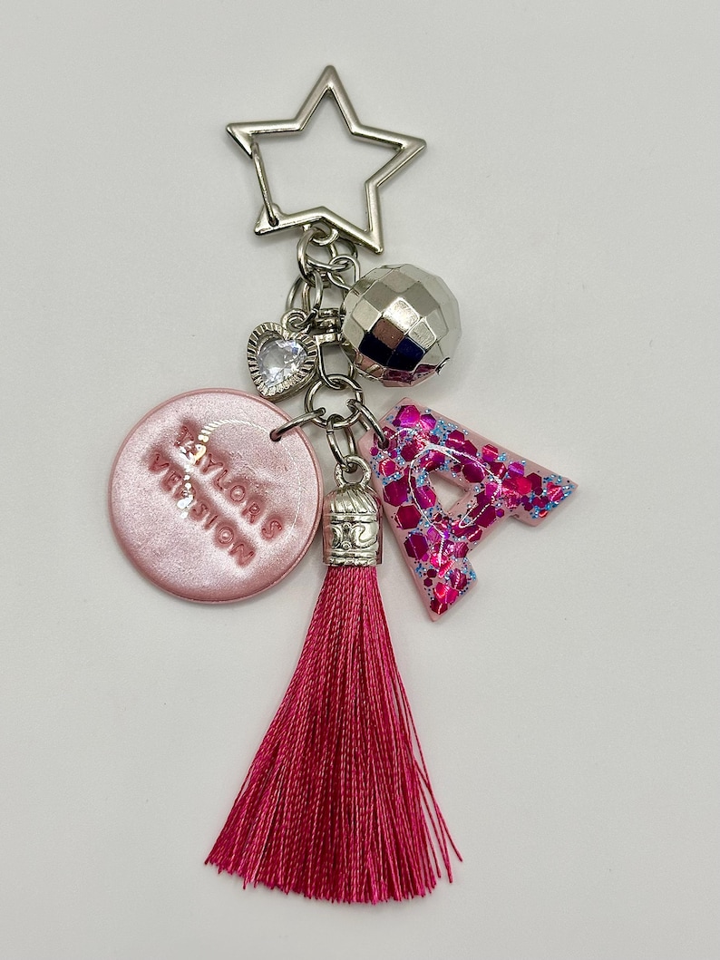 May include: A keychain with a silver star-shaped clasp, a disco ball charm, a heart charm, a pink circular charm with text, a pink glittery letter "A", and a long pink tassel. The keychain is a decorative accessory.