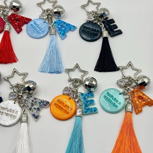 May include: A collection of keychains with star-shaped charms, disco ball accents, and colorful tassels. Each keychain features a letter or a circular pendant with the text "Taylor's Version". The letters and pendants are in various colors, including blue, orange, and black.