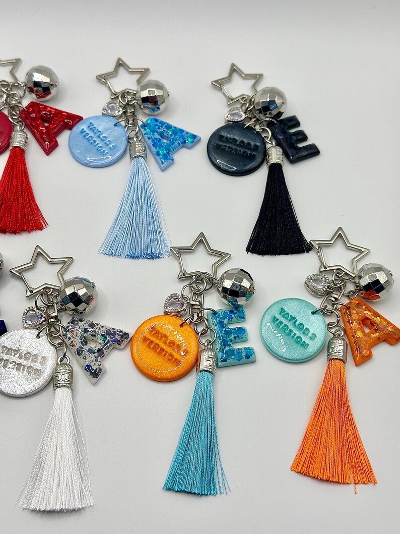 May include: A collection of keychains with star-shaped charms, disco ball accents, and colorful tassels. Each keychain features a letter or a circular pendant with the text "Taylor's Version". The letters and pendants are in various colors, including blue, orange, and black.