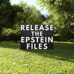 May include: A black yard sign with the text "RELEASE THE EPSTEIN FILES" in white block letters. The sign is placed in a grassy yard with trees in the background. The sign is held up by a metal wire frame.