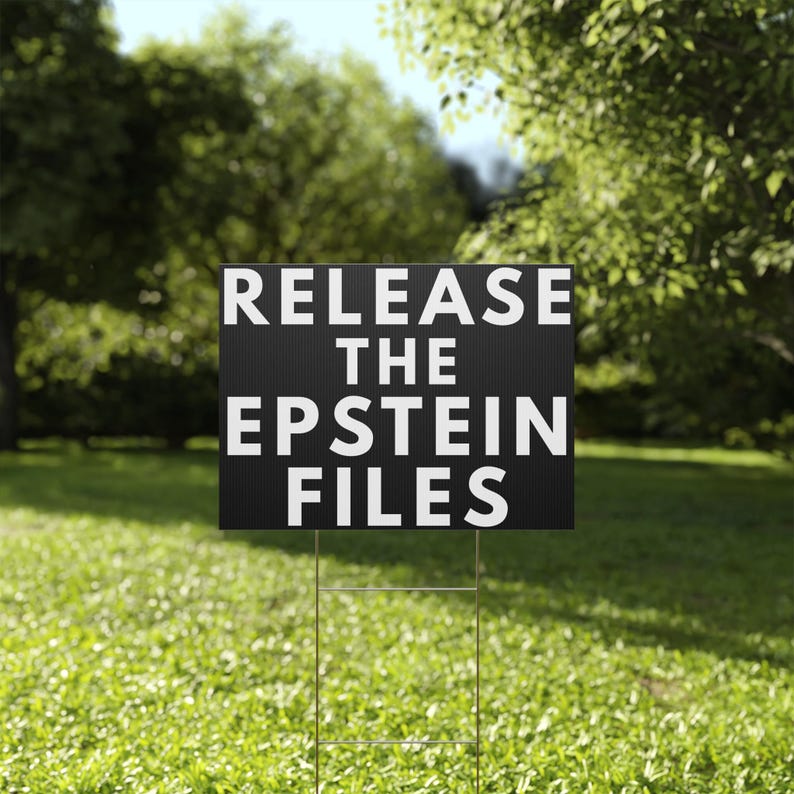 May include: A black yard sign with the text "RELEASE THE EPSTEIN FILES" in white block letters. The sign is placed in a grassy yard with trees in the background. The sign is held up by a metal wire frame.