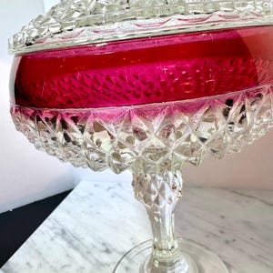 May include: A clear glass pedestal candy dish with a ruby red interior. The dish features a diamond-cut pattern on the base, stem, and lid. The lid is domed and fits snugly on the bowl.